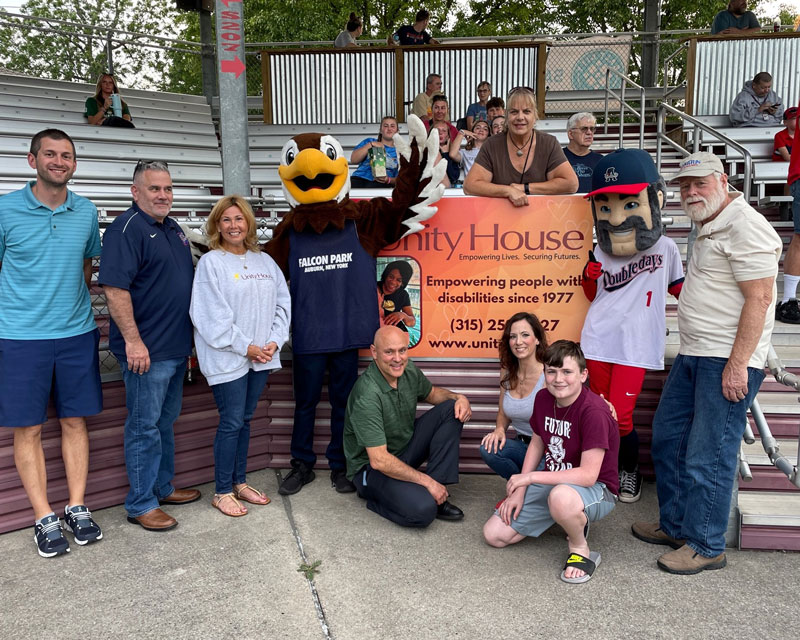 Group of Unity House Employees and Family Members at Falcon Park standing in front of a Unity House banner with Falcon Mascot.