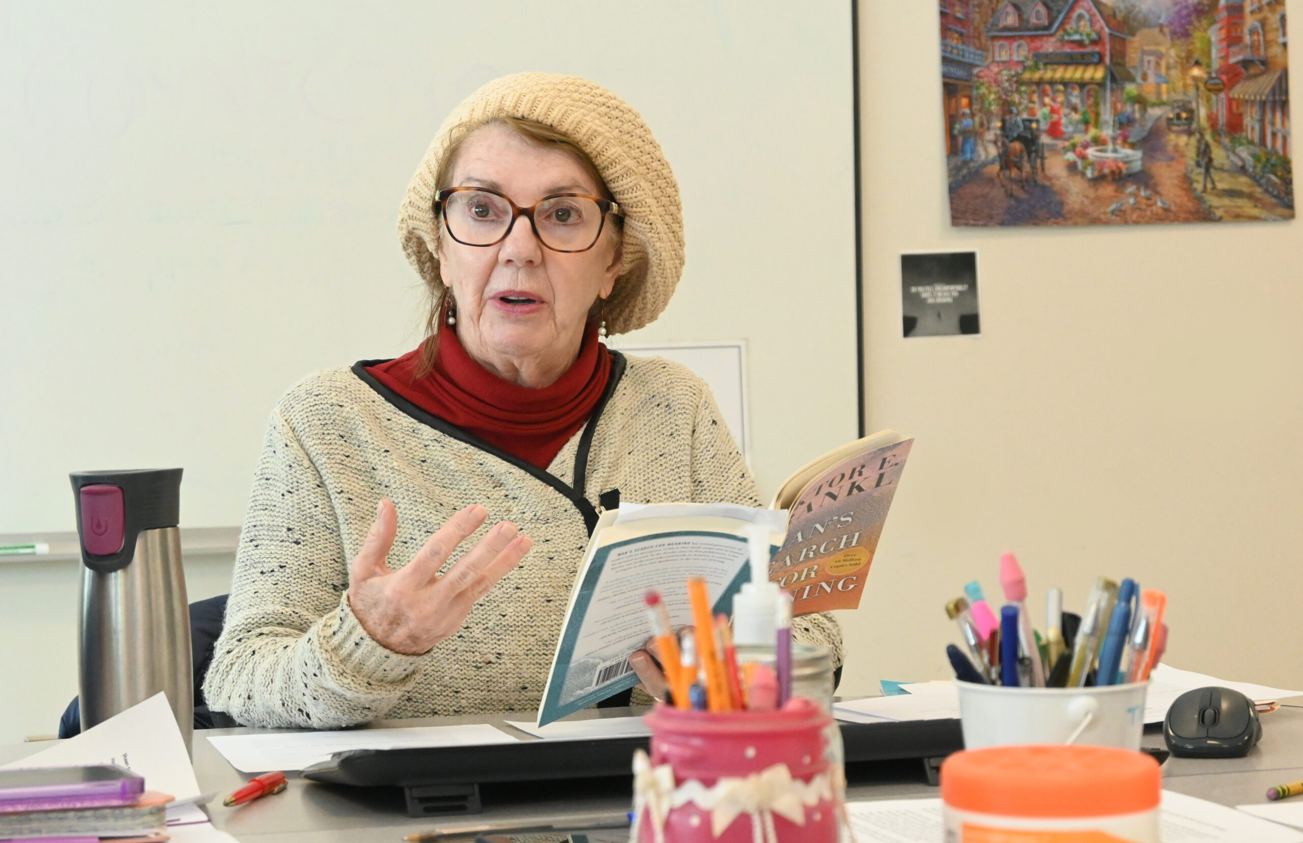 Elaine Meyers leads a creative writing class, holding a copy of Viktor E. Frankl's Man's Search for Meaning as she gestures passionately. The table in front of her is filled with colorful pens, papers, and a coffee mug, creating a welcoming classroom atmosphere.