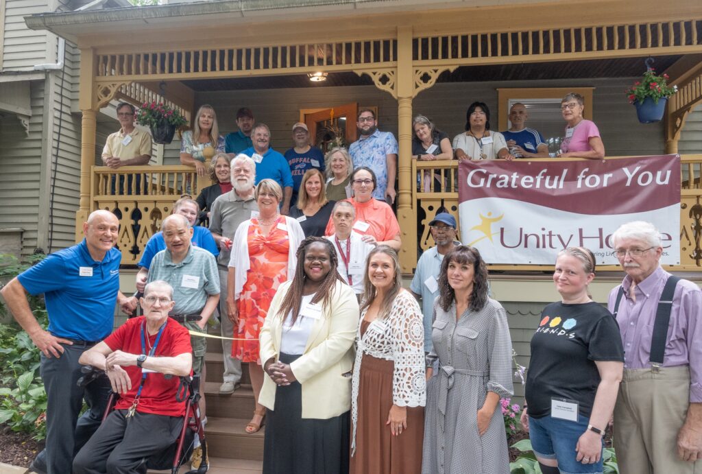 A diverse group of people pose together on the W Green St residence porch with a “Grateful for You” Unity House banner displayed. Some are standing, and some are sitting, all smiling at the camera.