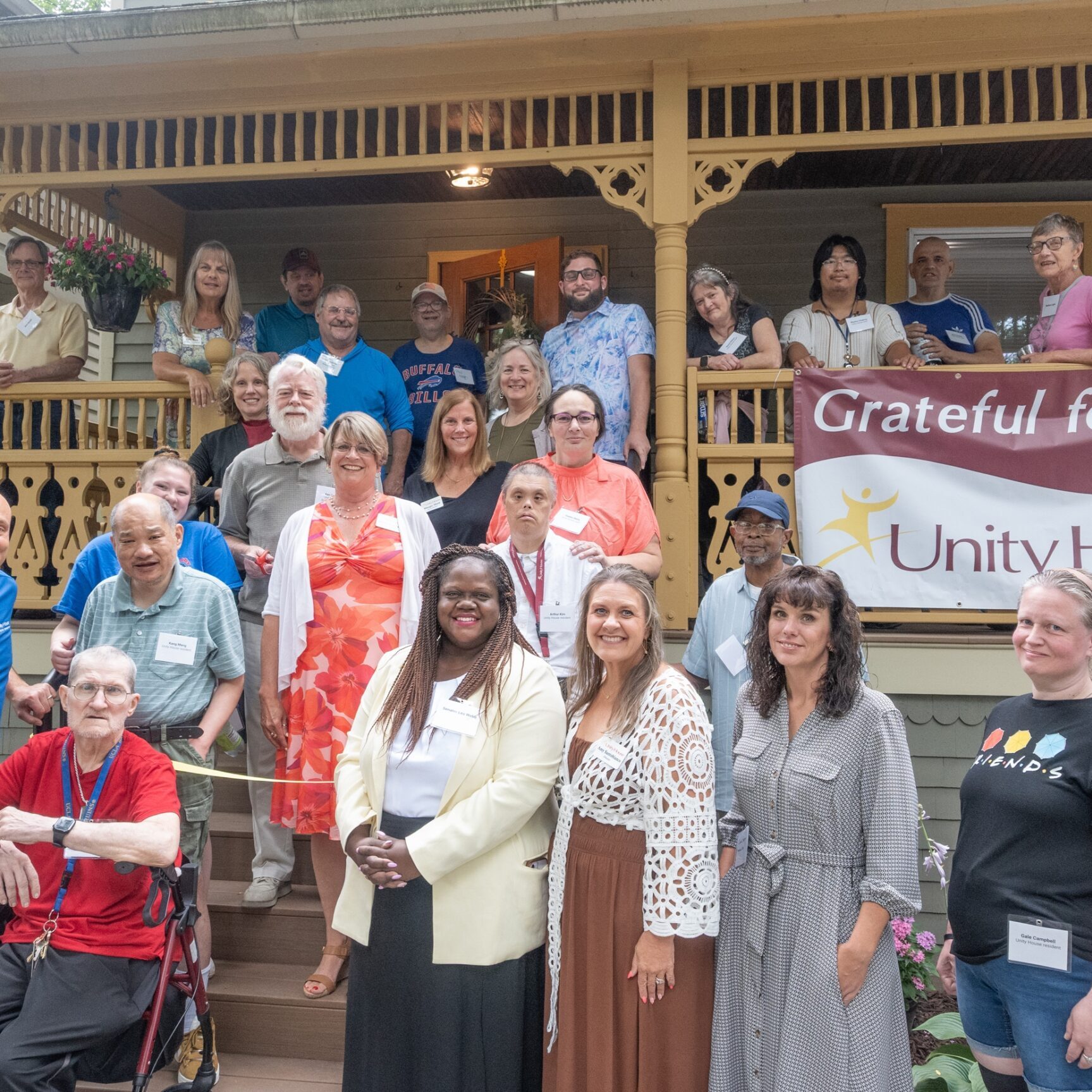 A diverse group of people pose together on the W Green St residence porch with a “Grateful for You” Unity House banner displayed. Some are standing, and some are sitting, all smiling at the camera.